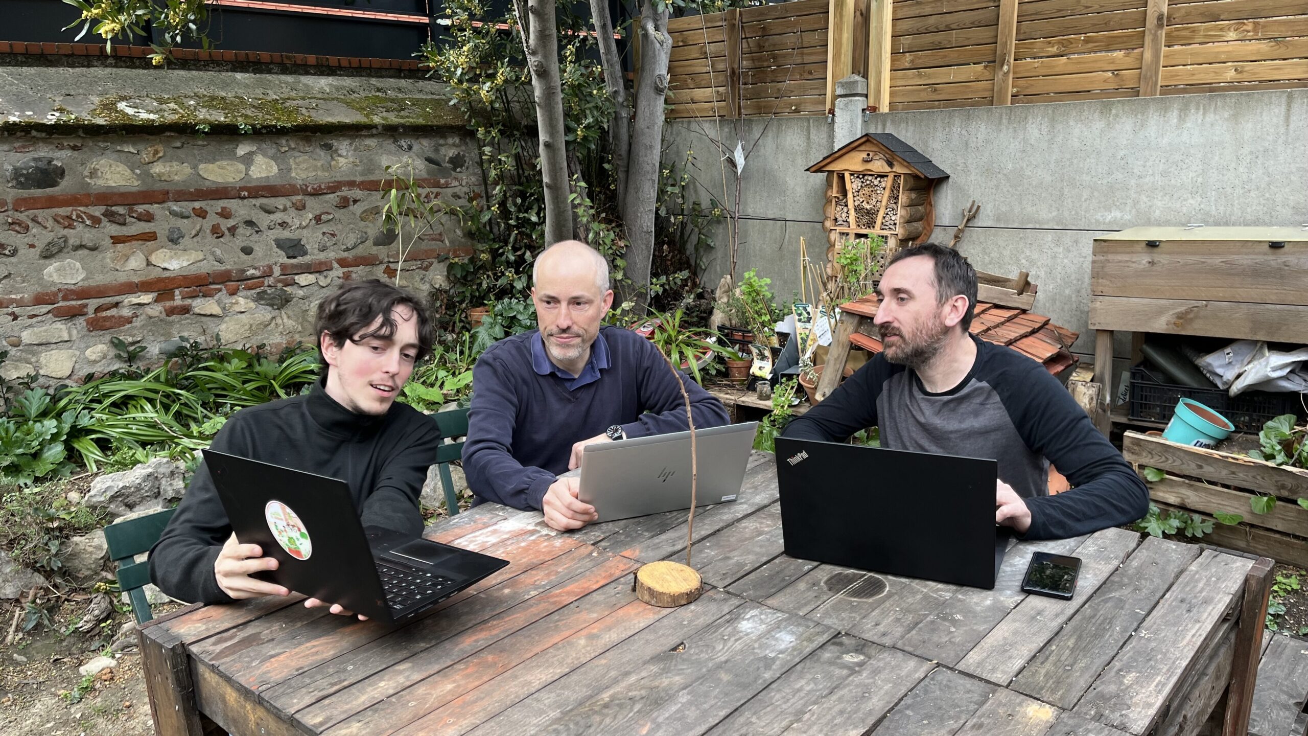 3 personnes travaillants sur une terrasse extérieur sur leurs ordinateurs. L'un montre aux autres son écran. Ils sont tous les 3 souriants